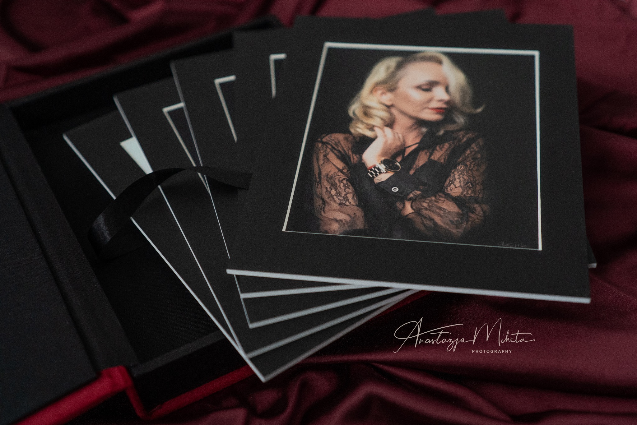 Elegancki zestaw portretów kobiecych w czarnych ramkach, ułożonych w pudełku z bordową wstążką, na tle bordowej tkaniny. Subtelne oświetlenie podkreśla detale.