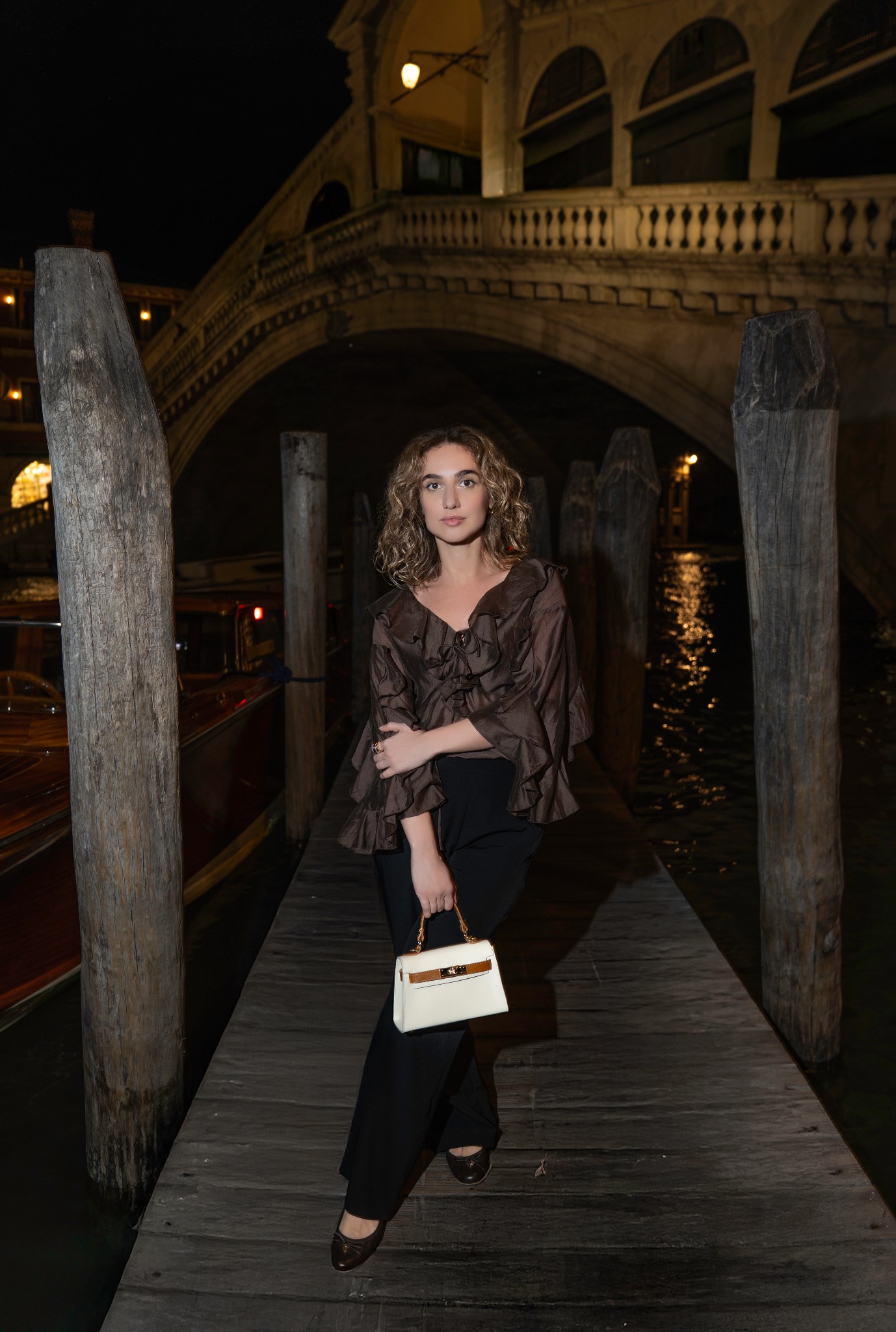 Stylowa sesja kobieca w Wenecji nocą: kobieta w brązowej bluzce i czarnych spodniach pozuje na drewnianym pomoście z mostem Rialto w tle. Elegancka torebka.