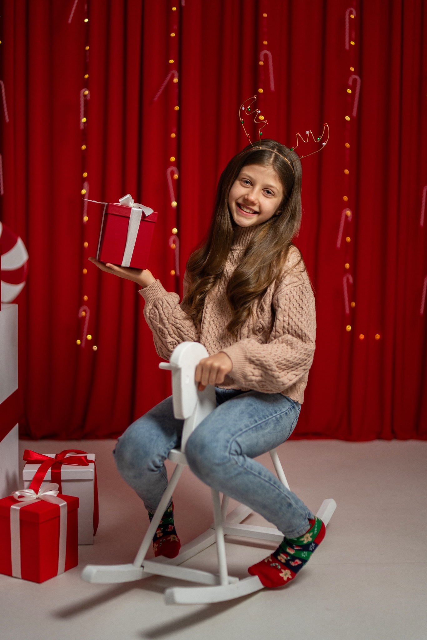 Uśmiechnięta dziewczynka w swetrze i jeansach siedzi na białym koniku na biegunach, trzymając czerwony prezent na tle czerwonej zasłony z lampkami.