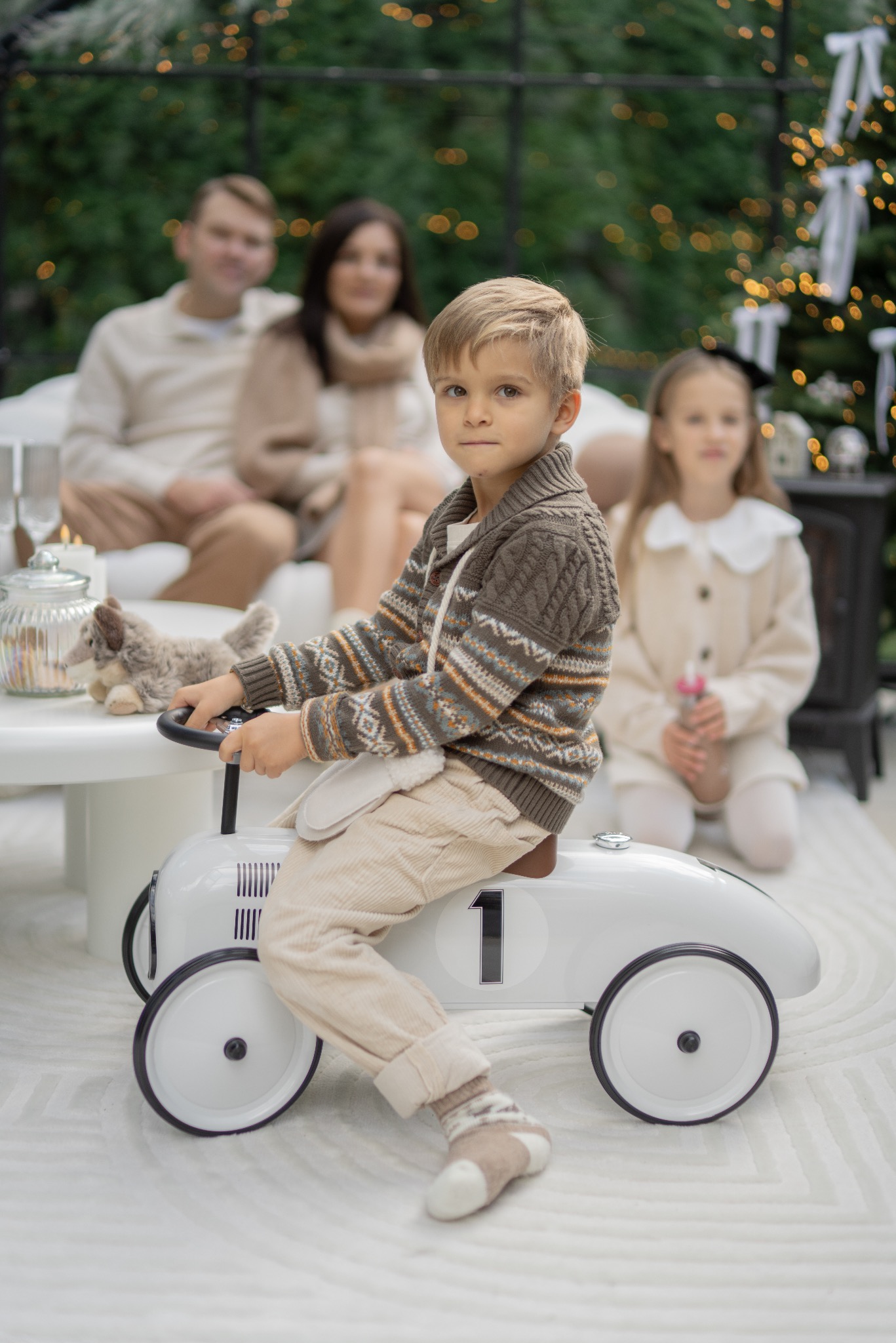 Chłopiec w swetrze siedzi na białym samochodzie-zabawce z numerem 1, w tle rodzina i choinka z lampkami. Ujęcie z sesji w neutralnej tonacji.