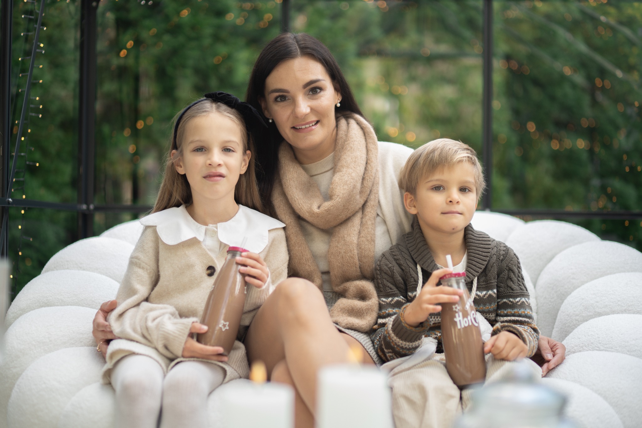 Portret rodzinny: mama z córką i synem siedzą na białej sofie, trzymając butelki z czekoladą. W tle zielona roślinność i delikatne światełka.