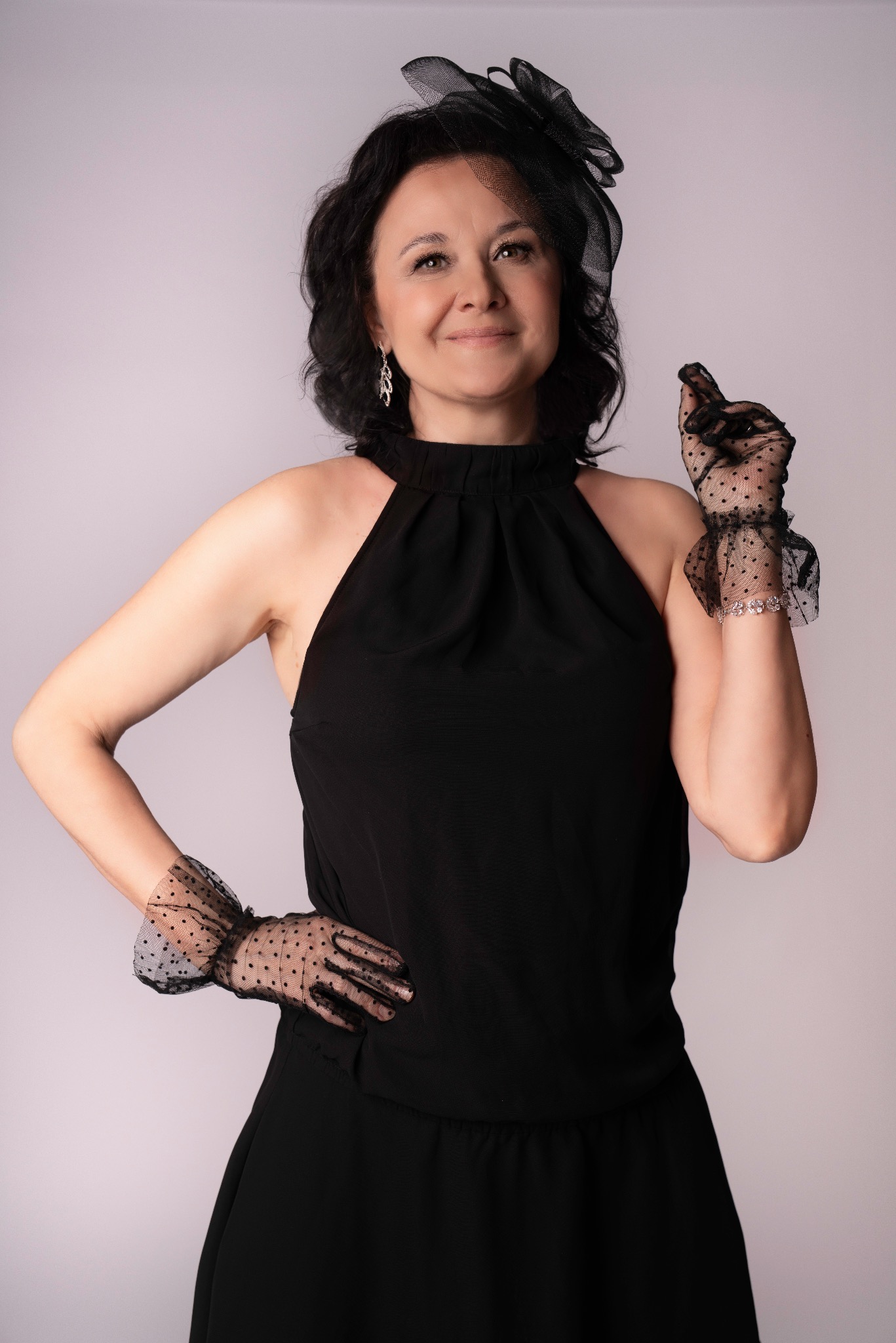 Elegancka kobieta w czarnej sukience i rękawiczkach z tiulu, z ozdobą na głowie, pozuje na jasnym tle. Uśmiech, subtelny makijaż, sesja w stylu retro.