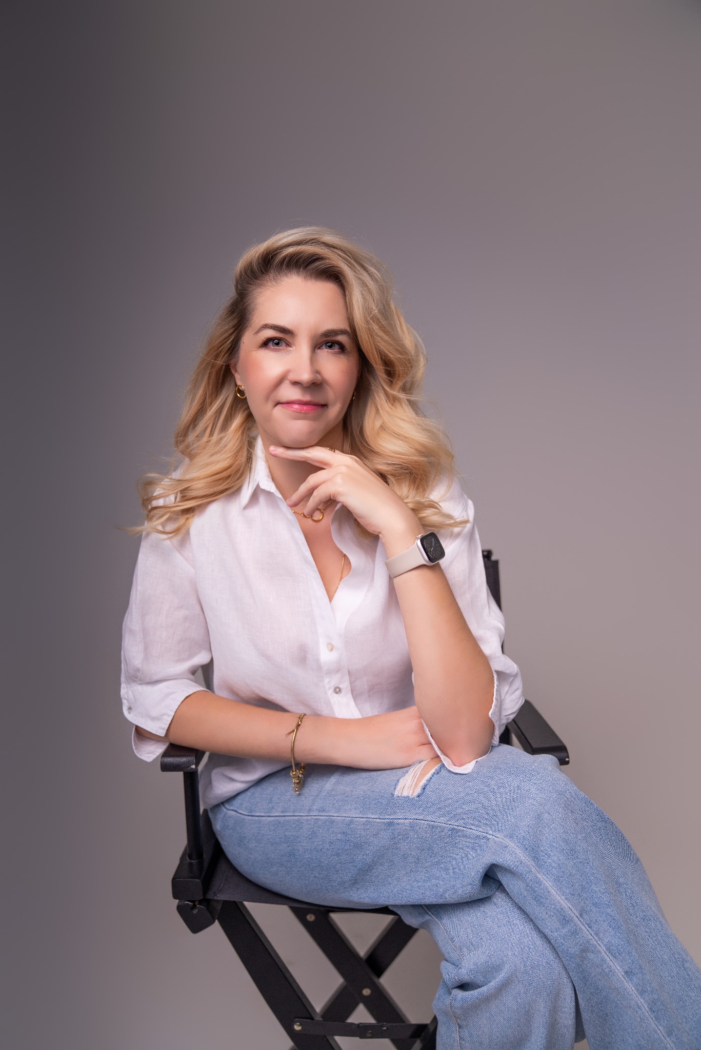 Portret kobiety z blond włosami, ubranej w białą koszulę i jeansy, siedzącej na czarnym krześle reżyserskim na szarym tle. Delikatny uśmiech, naturalny makijaż, złota biżuteria.