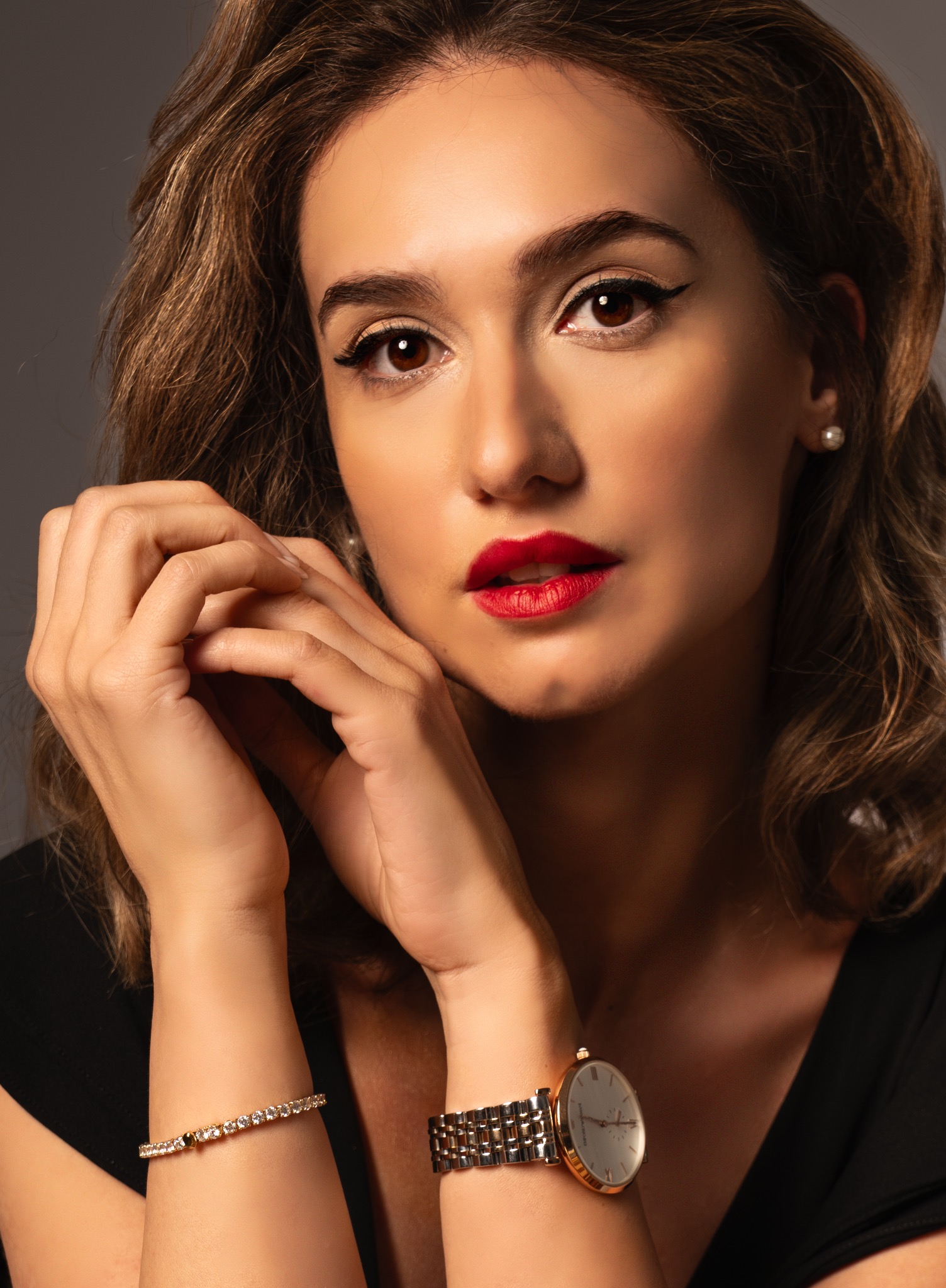 Portret kobiety z wyrazistym makijażem, dłońmi blisko twarzy, w eleganckiej biżuterii i zegarku. Ujęcie z bliska, podkreślające detale urody i akcesoriów.