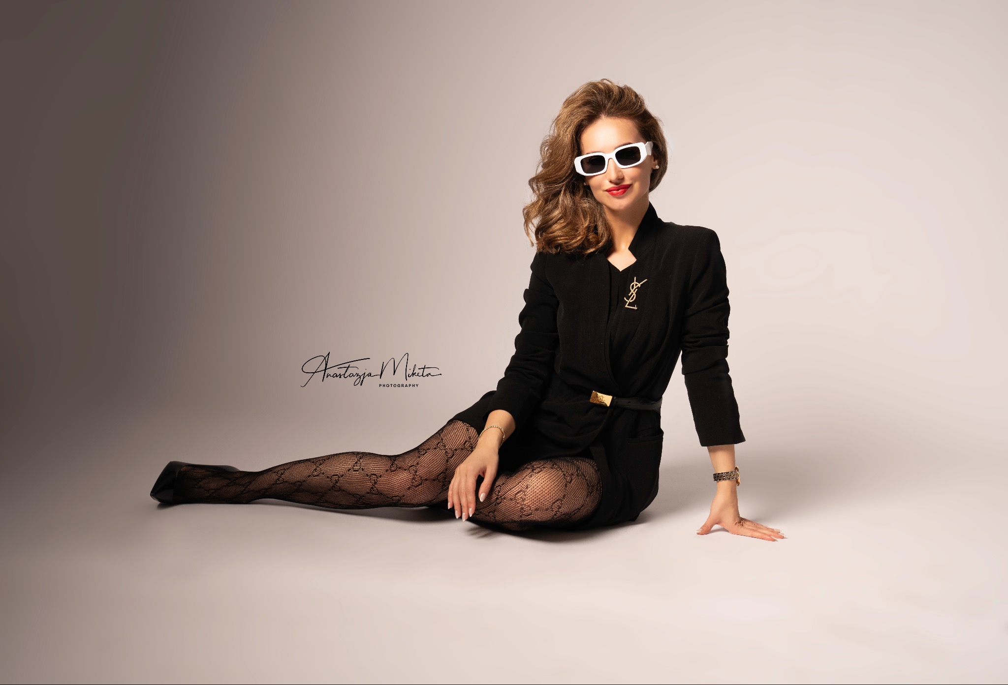 Stylowa kobieta w czarnej marynarce, wzorzystych rajstopach i białych okularach pozuje siedząc na podłodze w studio. Uwagę przykuwa złota broszka i pasek.