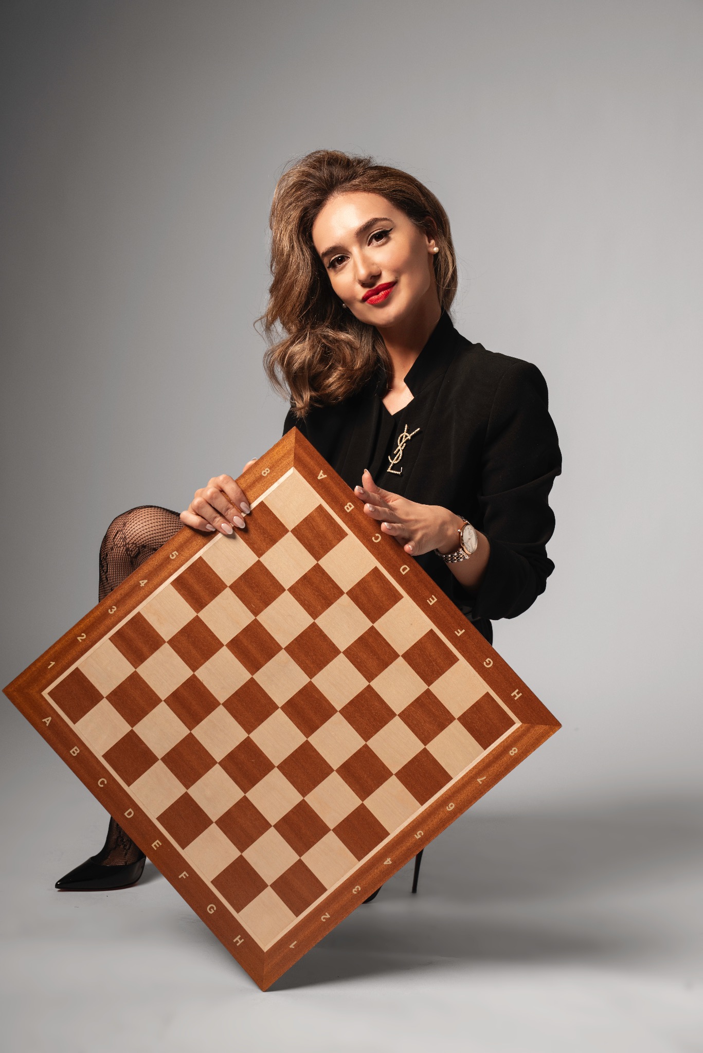 Stylowa kobieta w czarnej marynarce pozuje z szachownicą, prezentując makijaż z czerwoną szminką i delikatną biżuterię na szarym tle.