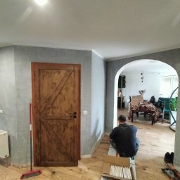 Tomasz Marciniak - Remont wnętrza: mężczyzna układa płytki podłogowe w pomieszczeniu z drewnianą podłogą i szarą ścianą. Widoczna drewniana stolarka drzwiowa i łukowe przejście.