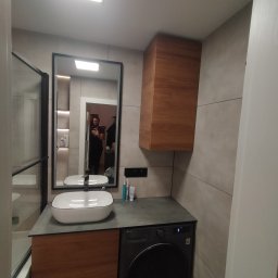 Tomasz Marciniak - Nowoczesna łazienka z umywalką nablatową, szafką z drewnianym frontem i pralką. W lustrze odbija się osoba robiąca zdjęcie. Minimalistyczny design.