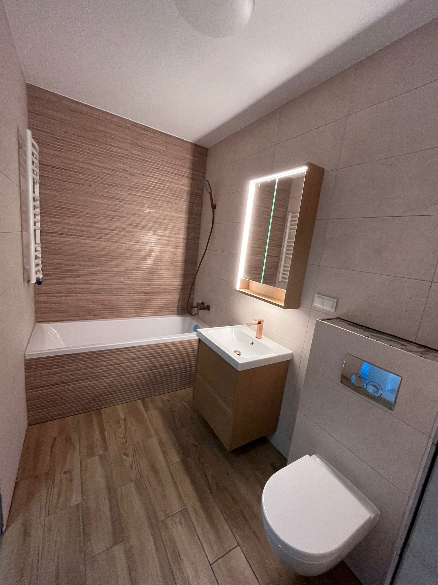 Wykończona łazienka w beżach i brązach: drewnopodobne płytki, podświetlane lustro, wanna obudowana płytkami, wisząca toaleta. Nowoczesny design.