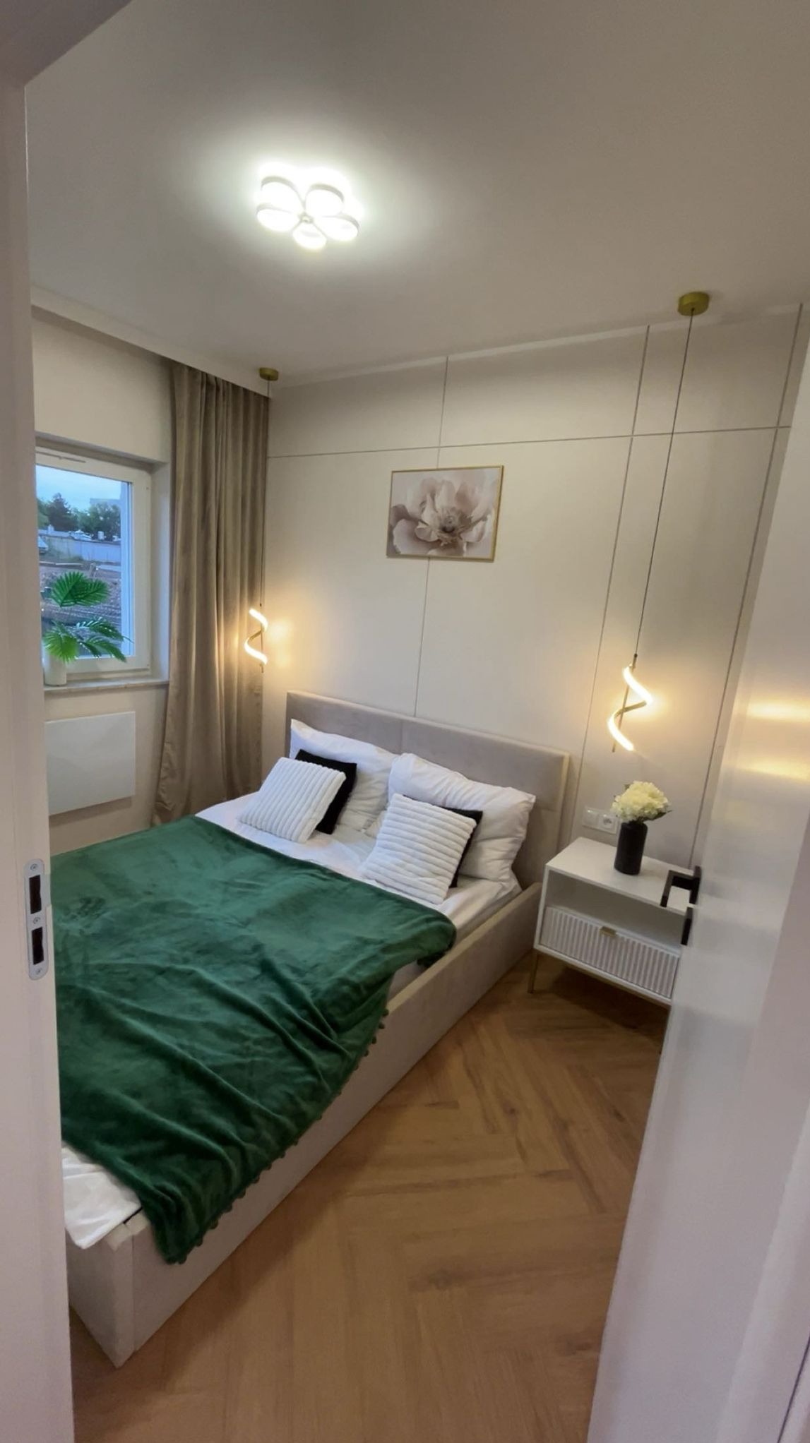 Nowoczesna sypialnia z zieloną narzutą na łóżku, parkietem w jodełkę i minimalistycznym oświetleniem. Na ścianie obraz z motywem kwiatowym.