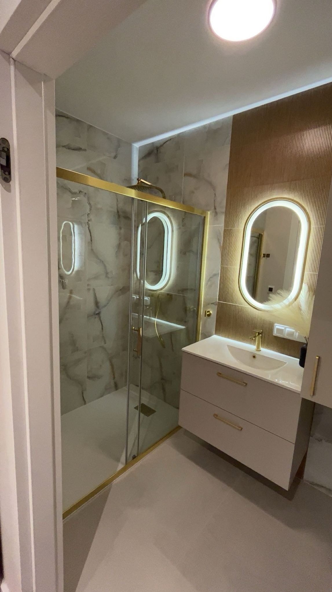 Nowoczesna łazienka po remoncie: kabina prysznicowa ze złotymi detalami, umywalka z szafką i podświetlane lustro owalne na tle marmurowych płytek.