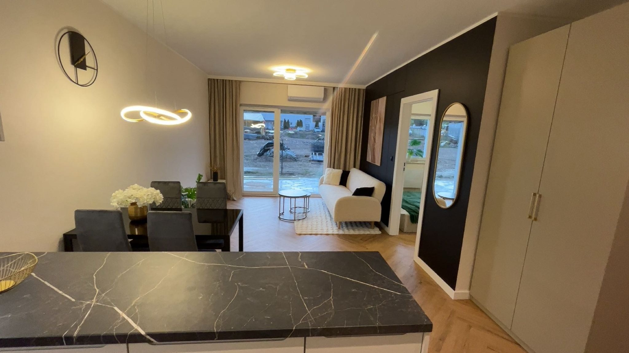 Nowoczesne wnętrze: czarny blat kuchenny, stół z krzesłami, sofa przy oknie balkonowym, czarna ściana z lustrem i szafa w jasnym odcieniu.