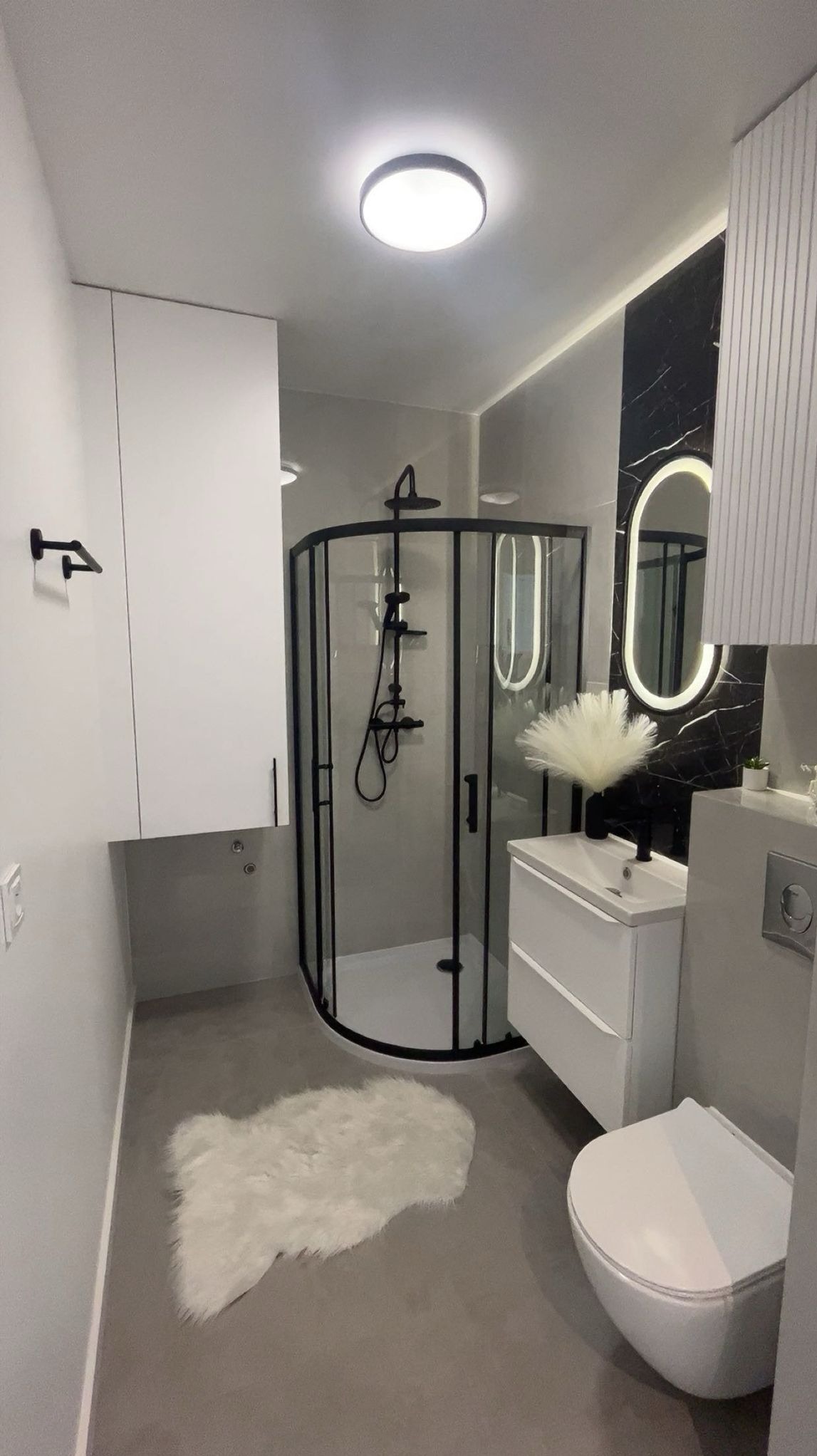 Nowoczesna łazienka z kabiną prysznicową w czarnej ramie, białą umywalką i podświetlanym lustrem. Szary odcień ścian i podłogi, akcent czarnego marmuru.
