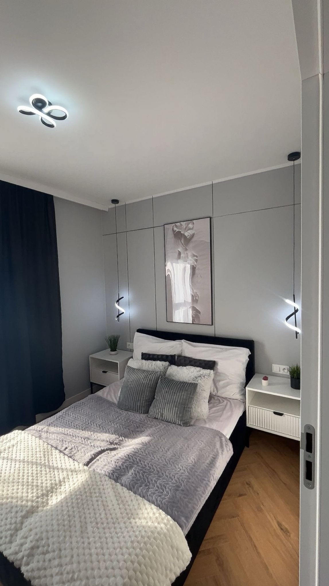 Nowoczesna sypialnia w szarościach z drewnianą podłogą. Minimalistyczne lampy wiszące i geometryczne panele ścienne. Biała pościel i dekoracyjne poduszki.