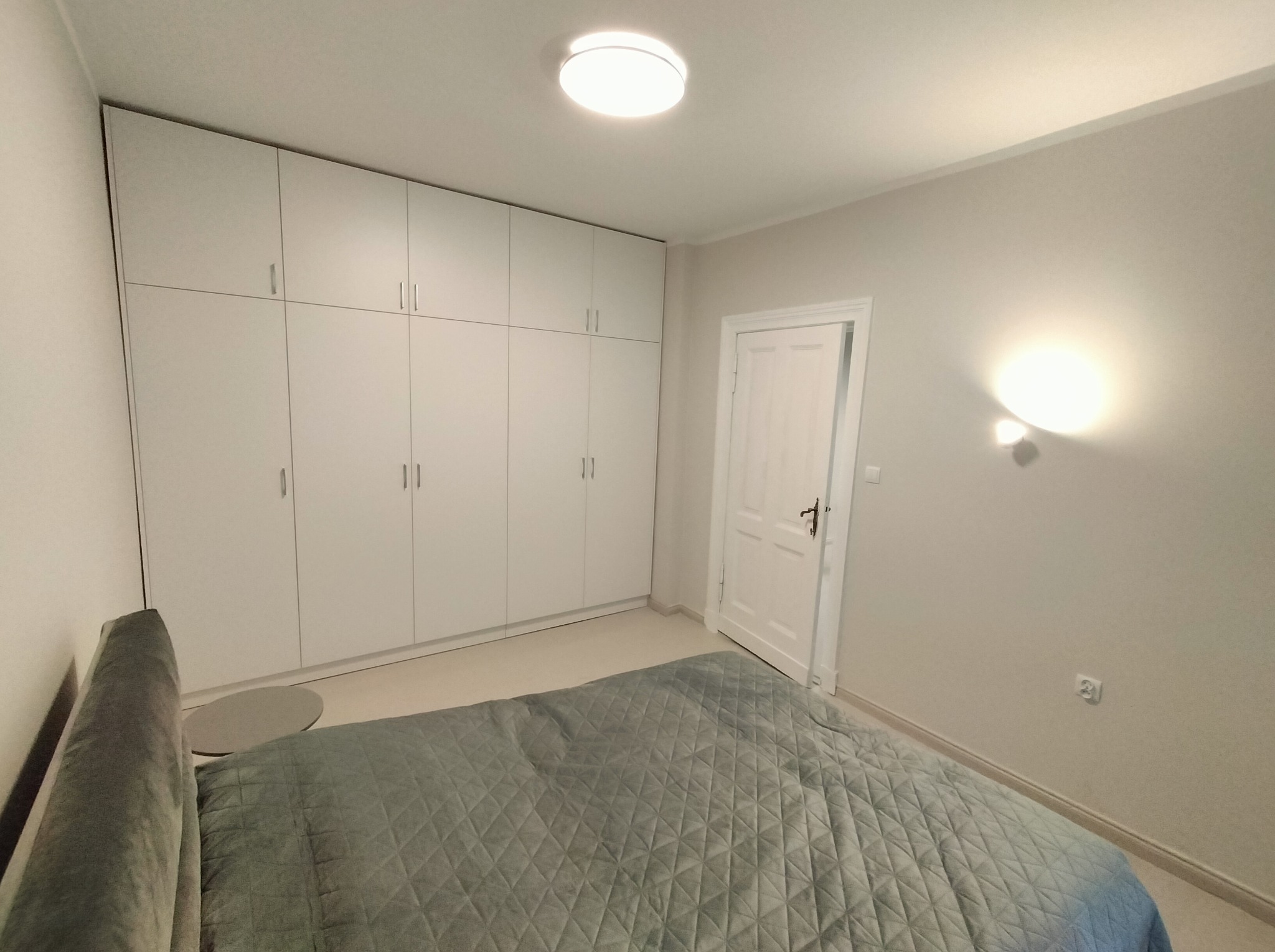 Jasna sypialnia z dużym, białym, zabudowanym szafą, łóżkiem z szarozieloną narzutą i otwartymi białymi drzwiami. Minimalistyczny design, stonowana kolorystyka.