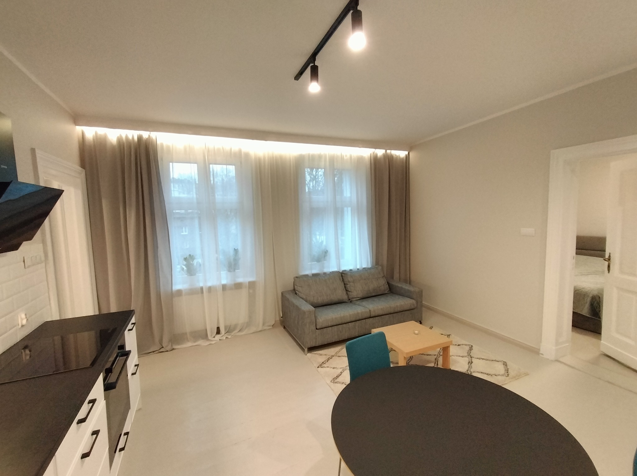 Jasne wnętrze po remoncie: salon z sofą, stolikiem kawowym i jadalnią z okrągłym stołem. Widok na sypialnię przez otwarte drzwi. Minimalistyczny design, neutralna kolorystyka.