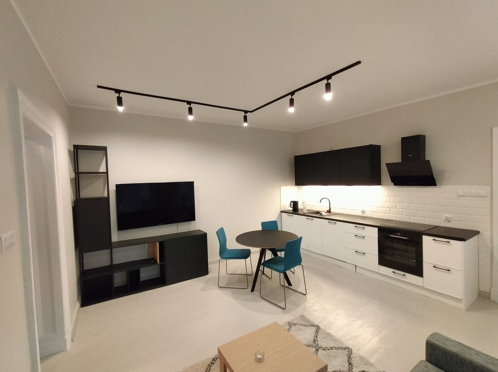 Nowoczesne wnętrze po remoncie: biała kuchnia z czarnymi akcentami, stół z krzesłami, szafka RTV z telewizorem. Oświetlenie szynowe.