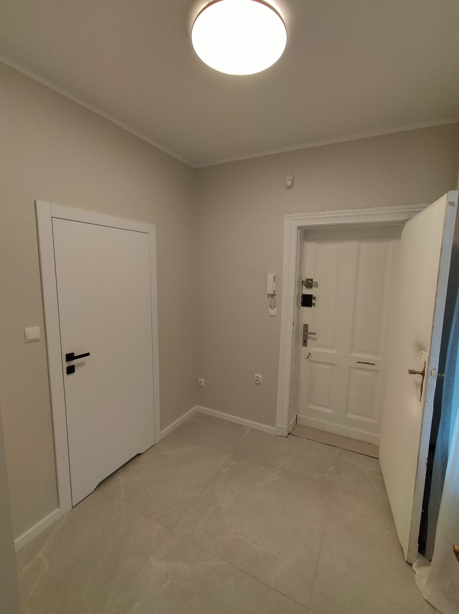 Jasne wnętrze po remoncie: dwie białe, nowoczesne drzwi z czarnymi klamkami, szara podłoga z płytek, neutralne ściany i sufit z okrągłą lampą.