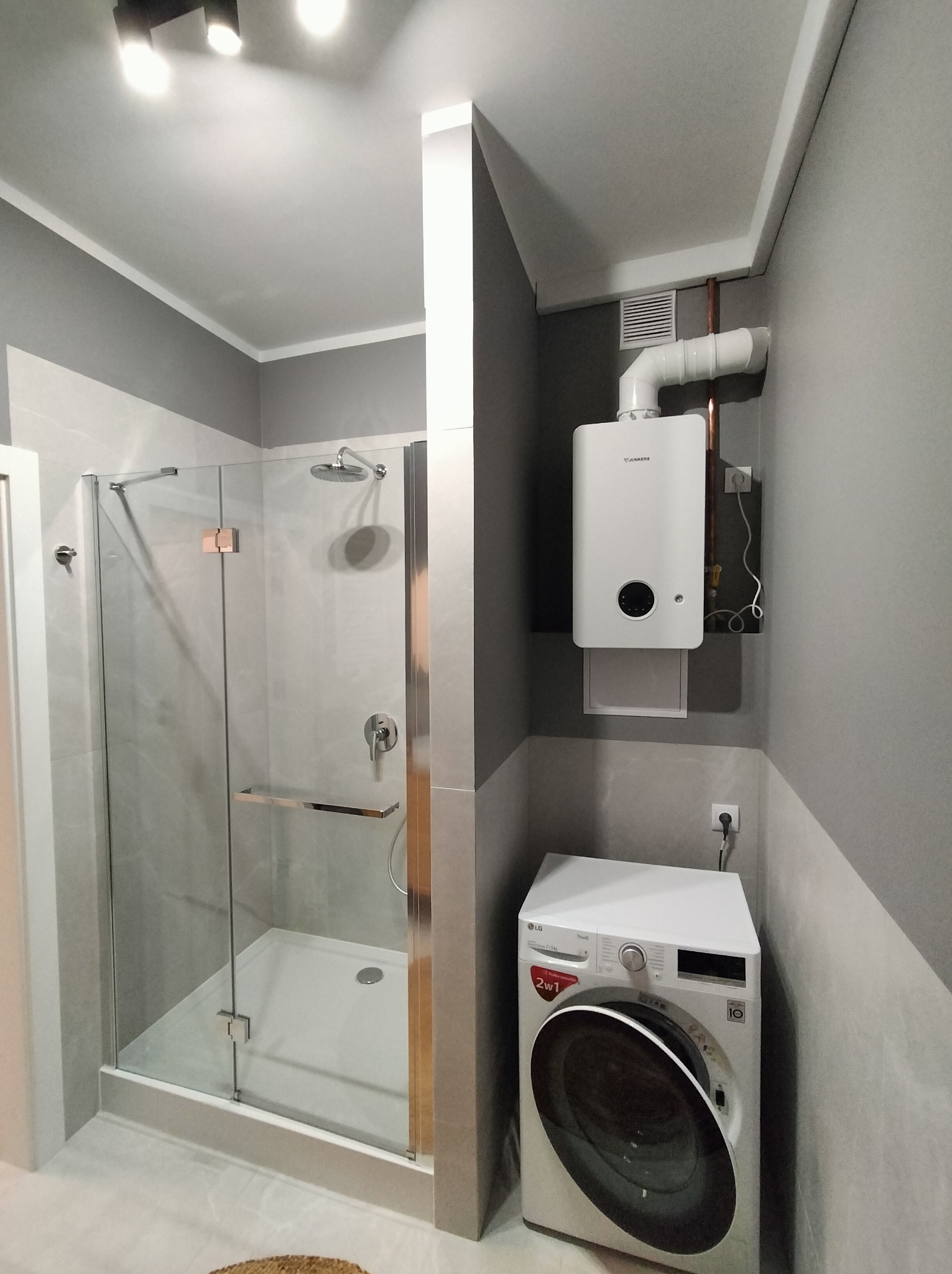 Nowoczesna łazienka w szarościach: kabina prysznicowa ze srebrnymi okuciami, biały piec gazowy Junkers i pralka LG 2w1. Minimalistyczny design, funkcjonalność i elegancja.