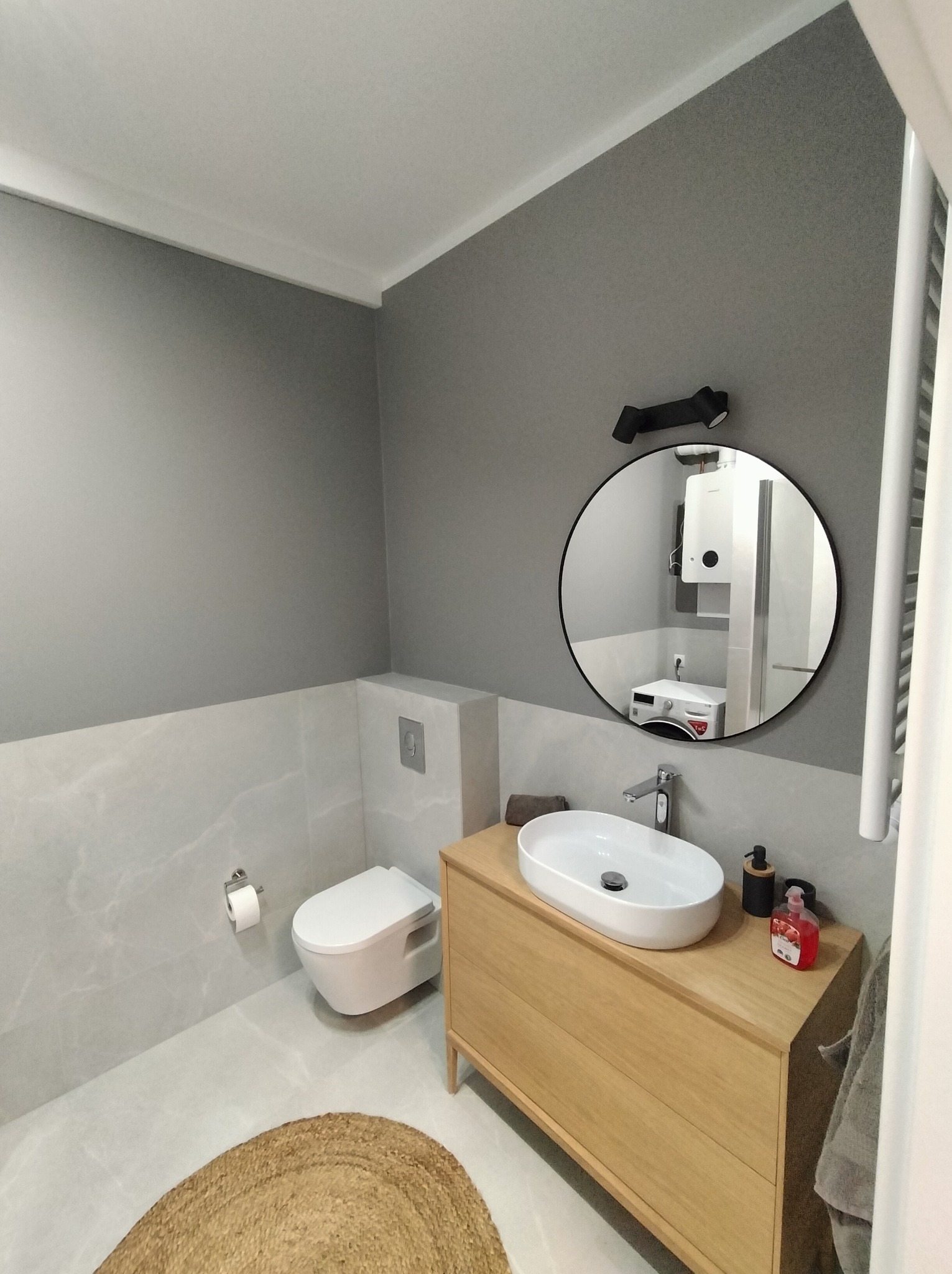 Nowoczesna łazienka w szarościach z drewnianą szafką pod umywalką i okrągłym lustrem, w którym odbija się pralka i bojler. Minimalistyczny design.