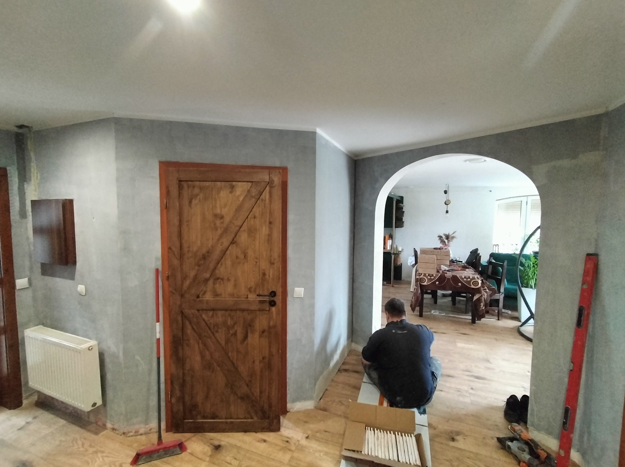 Remont wnętrza: mężczyzna układa płytki podłogowe w pomieszczeniu z drewnianą podłogą i szarą ścianą. Widoczna drewniana stolarka drzwiowa i łukowe przejście.