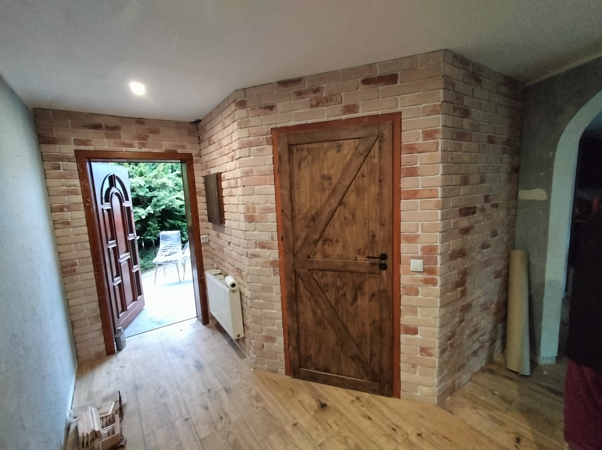 Wykończone wnętrze z ceglaną ścianą, drewnianą podłogą i drzwiami w stylu rustykalnym. Widoczny fragment grzejnika i otwarte drzwi na zewnątrz.