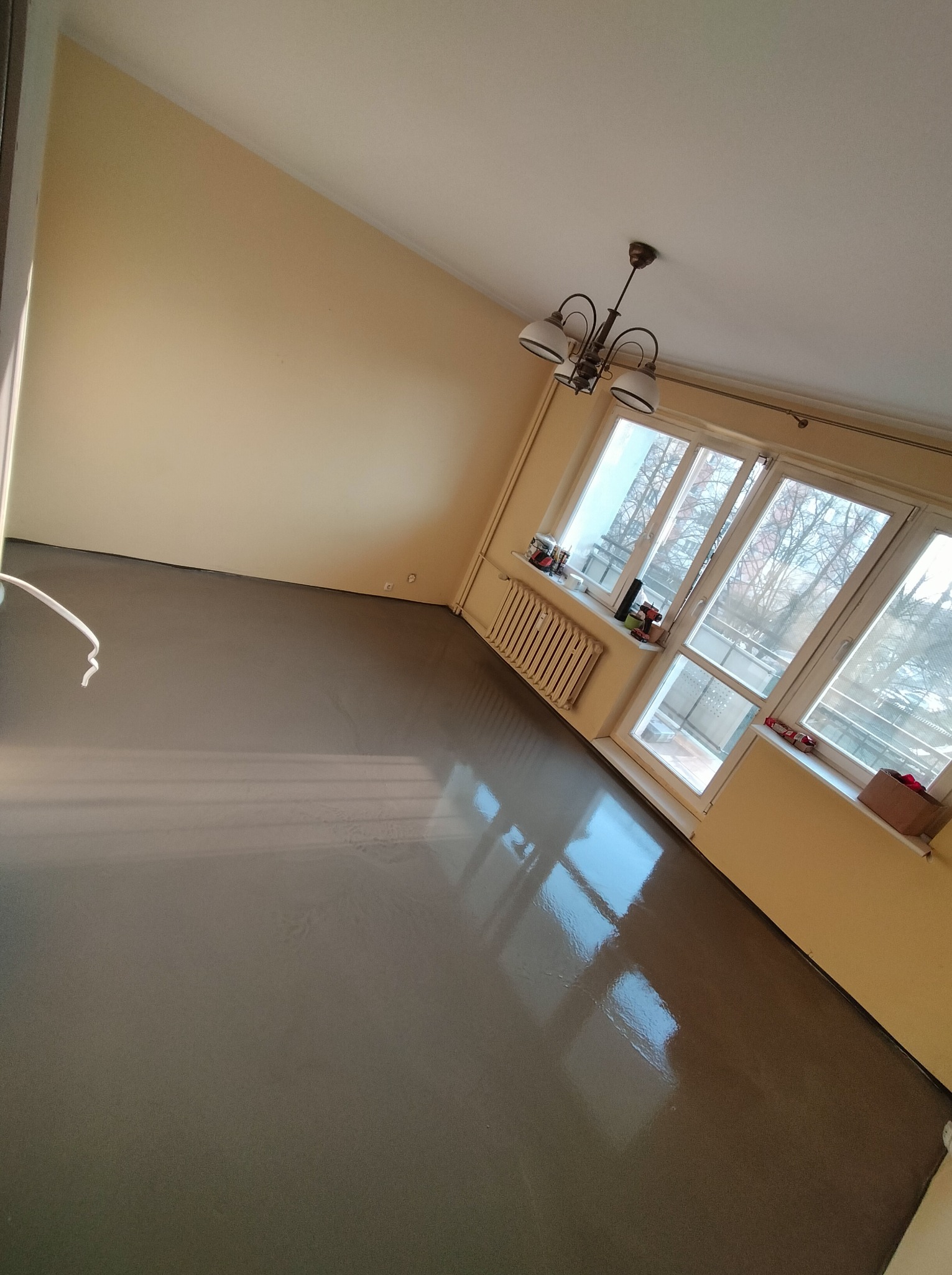 Wylany, gładki, samopoziomujący podkład podłogowy w pokoju z oknem balkonowym. Widoczny grzejnik i lampa sufitowa. Świeże wylewki podłogowe w odcieniach szarości.
