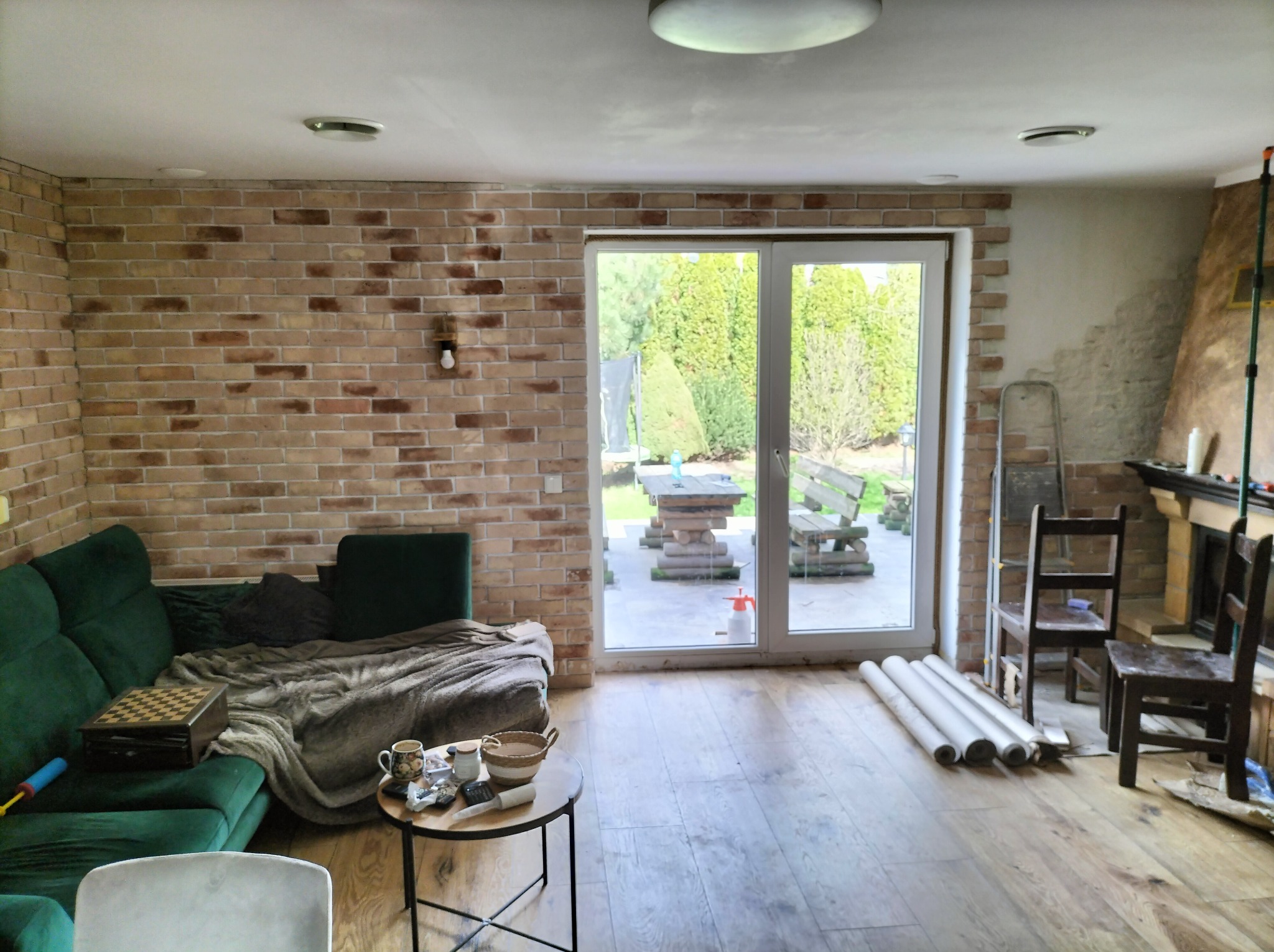 Salon w trakcie remontu: ceglana ściana, okno z widokiem na ogród, podłoga z paneli, sofa i narzędzia malarskie. Częściowo wykończone ściany.