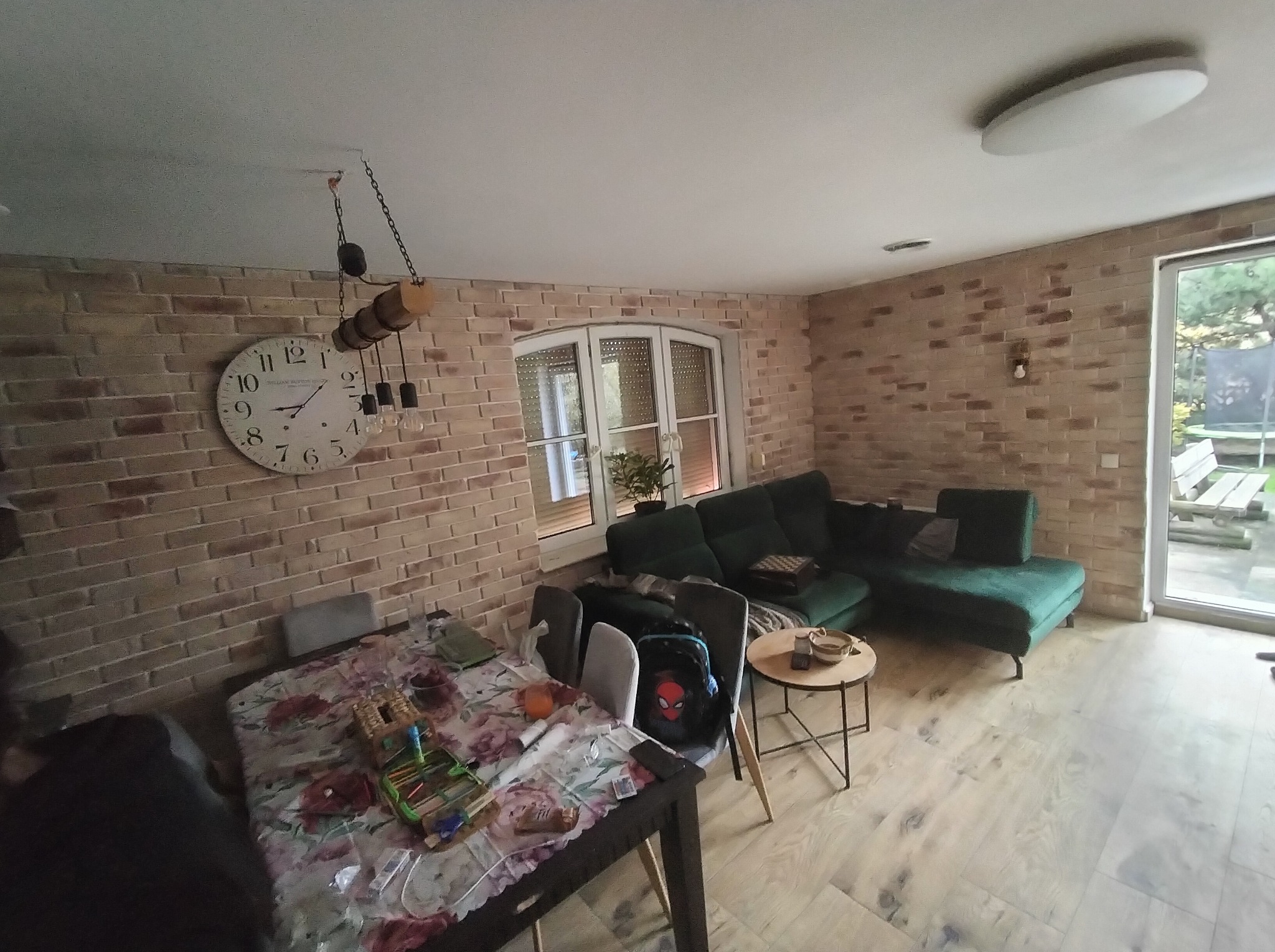 Salon z ceglanymi ścianami, zieloną sofą narożną i drewnianą podłogą. Na ścianie zegar i lampa wisząca. Widoczne okno i drzwi balkonowe.