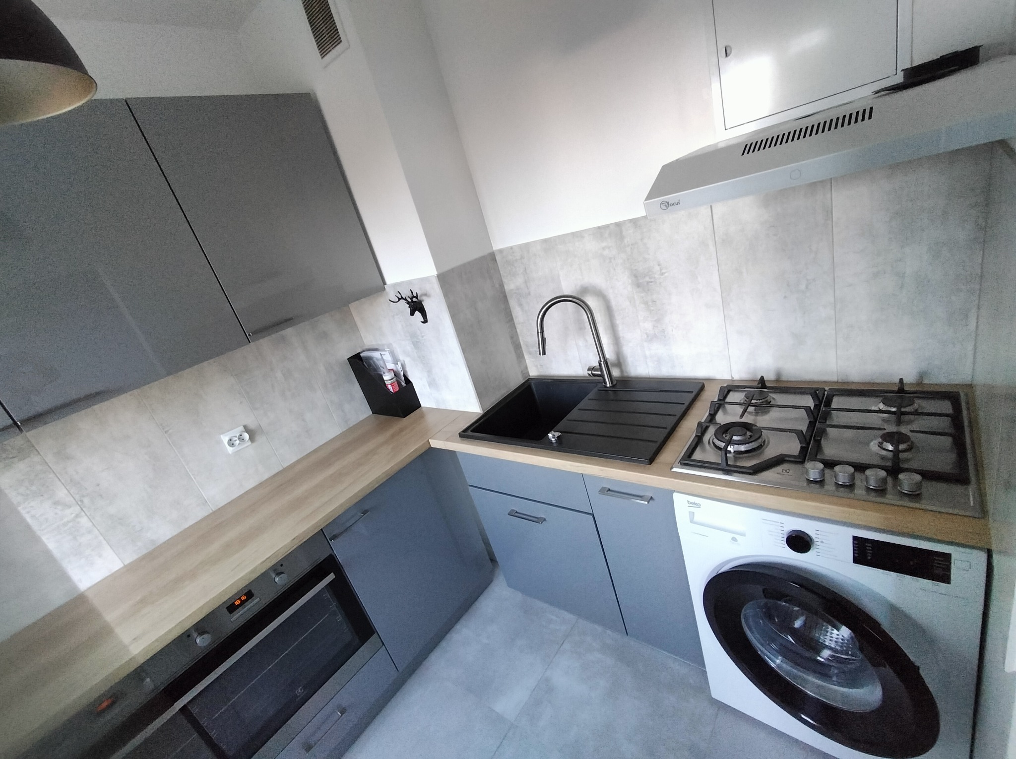 Nowoczesna kuchnia w szarościach: zabudowa z piekarnikiem, płyta gazowa, zlew i pralka w jednej linii. Minimalistyczny design, jasne blaty i płytki imitujące beton.