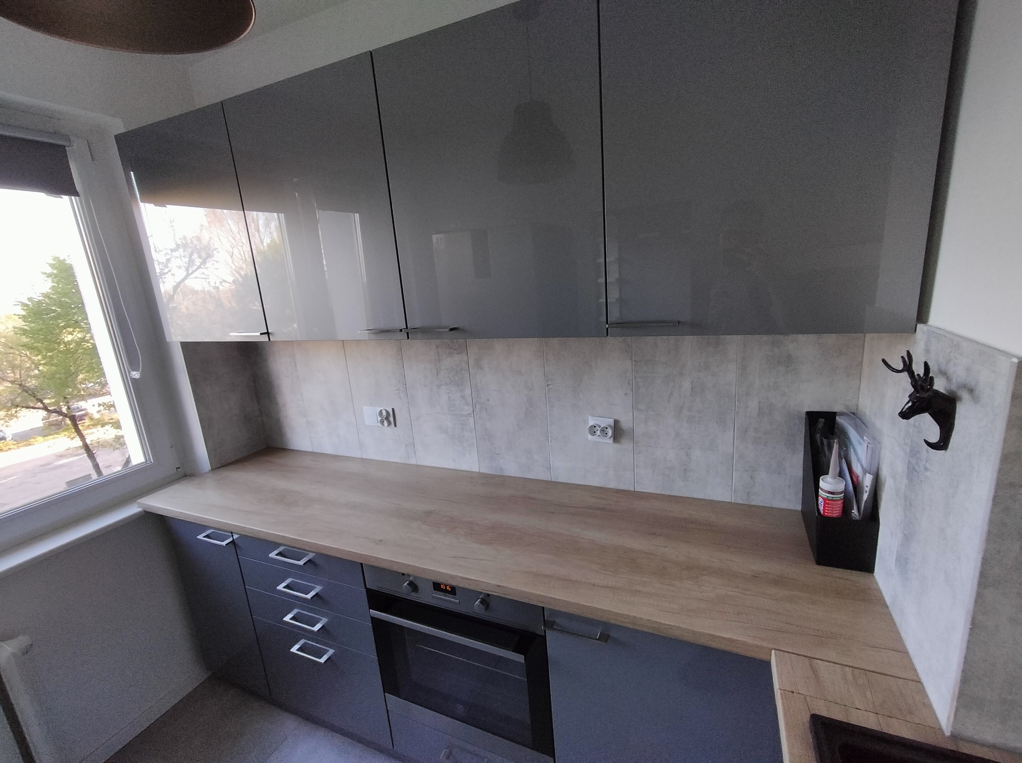 Szara kuchnia z drewnianym blatem i błyszczącymi szafkami górnymi. Widok na okno i dekoracyjny wieszak w kształcie głowy jelenia. Nowoczesny design.