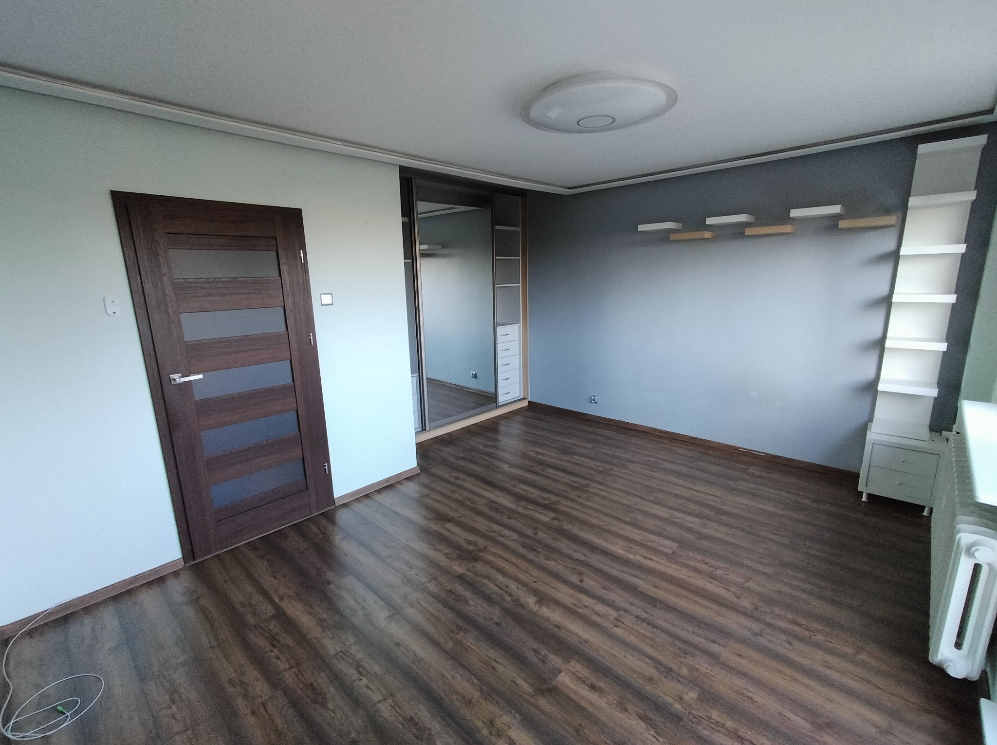 Pokój po remoncie: ciemna podłoga, szare ściany, brązowe drzwi z przeszkleniami, biała szafa wnękowa z lustrami i półki na ścianie. Nowoczesne wykończenie wnętrza.