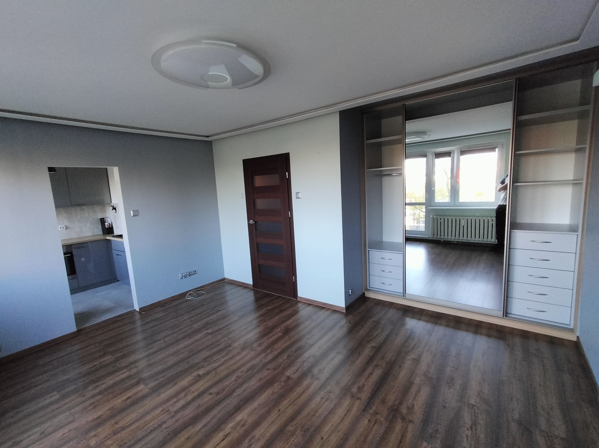 Pokój po remoncie: drewniana podłoga, zabudowana szafa z lustrami, widok na kuchnię. Świeżo wykończone wnętrze z nowoczesnym oświetleniem sufitowym.