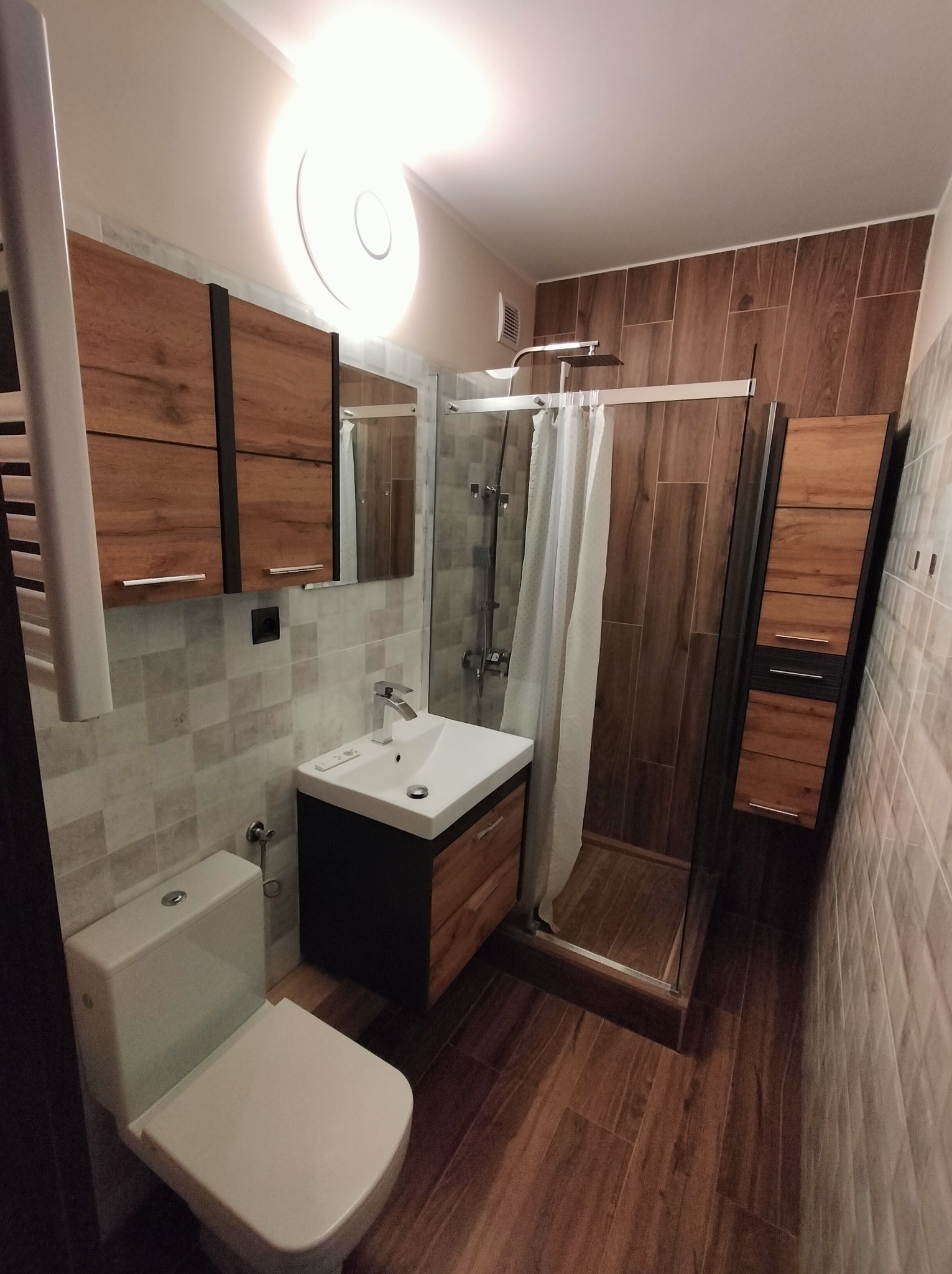 Nowoczesna łazienka po remoncie: kabina prysznicowa ze szklaną ścianką, umywalka z szafką, toaleta i grzejnik drabinkowy. Brązowe płytki imitujące drewno.
