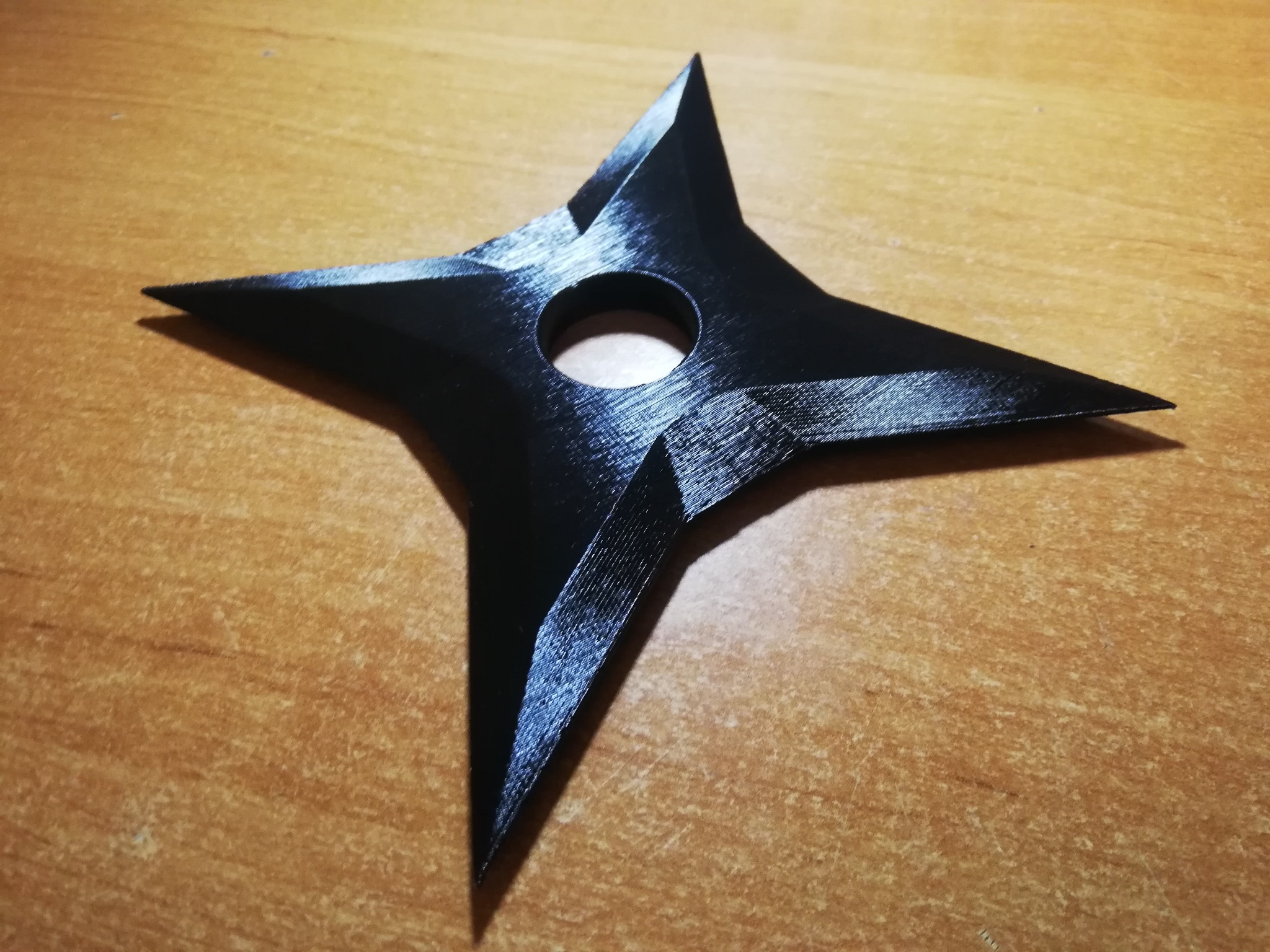 Czarny, czteroramienny shuriken z otworem pośrodku, wydrukowany z tworzywa sztucznego, leży na drewnianym blacie. Widoczna tekstura warstw.