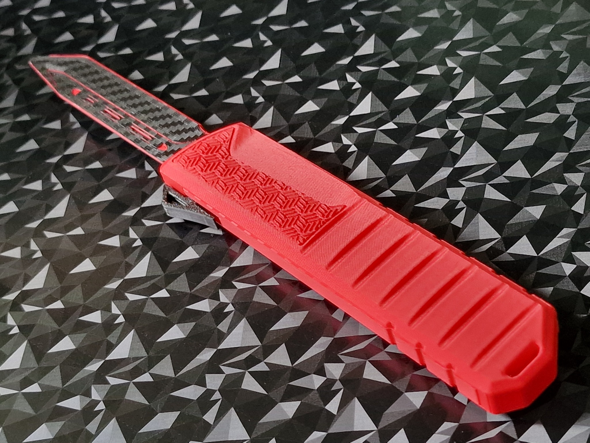 Czerwony nóż sprężynowy z ostrzem z włókna węglowego na tle geometrycznym. Widoczne detale tekstury i precyzja wykonania.