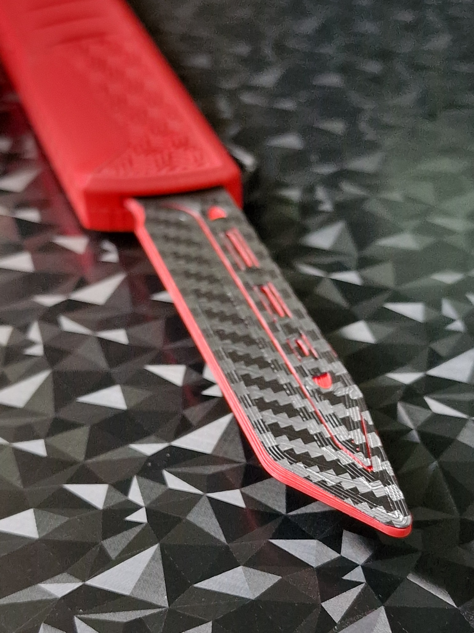 Czerwono-czarny element z wzorem włókna węglowego, prawdopodobnie wykonany metodą addytywną, leży na tle z geometrycznym wzorem trójkątów.
