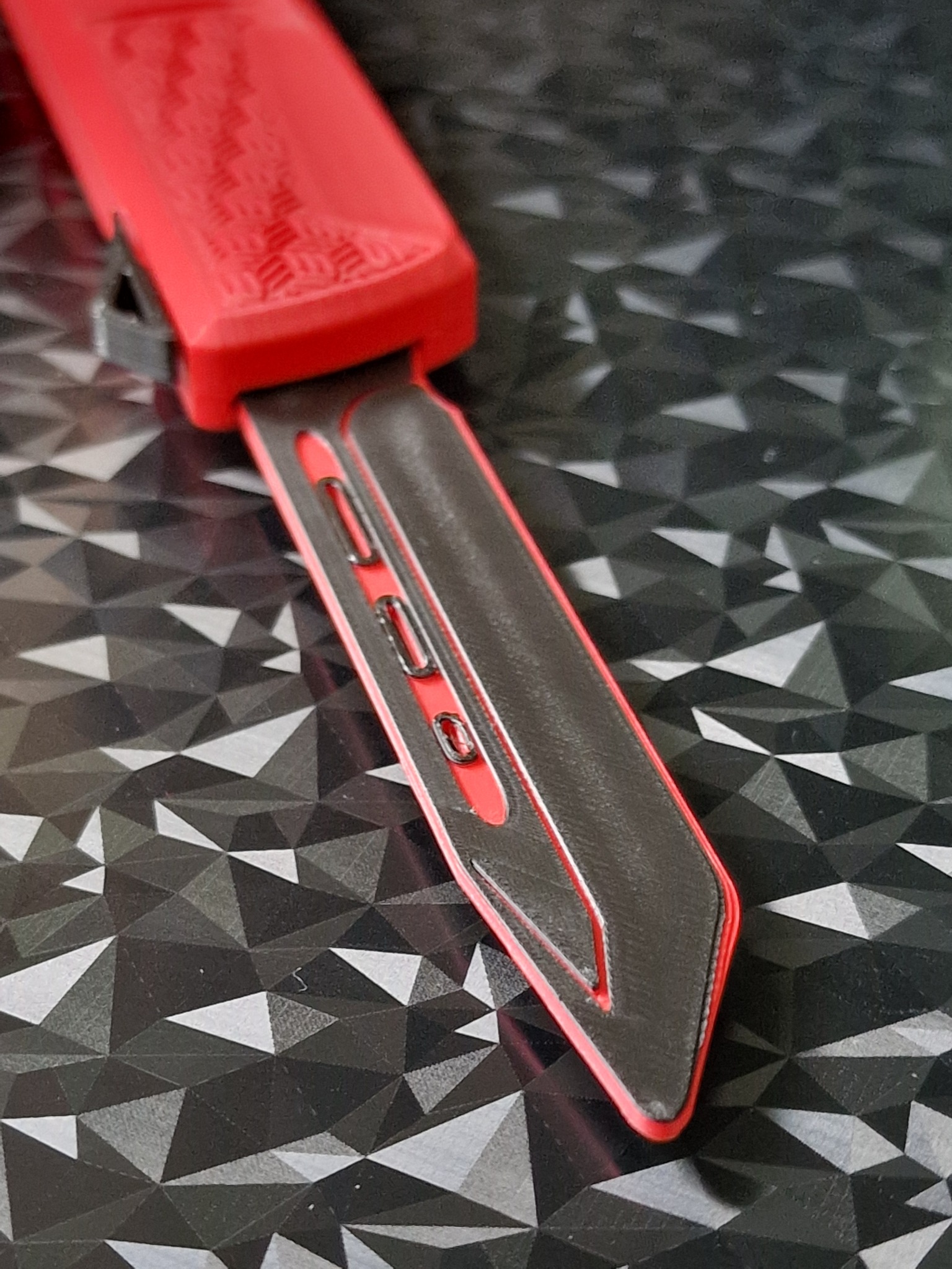 Czerwono-czarny, trójwymiarowy model klucza samochodowego wydrukowany na drukarce 3D, leżący na tle z geometrycznym wzorem. Widoczne detale i tekstura druku.