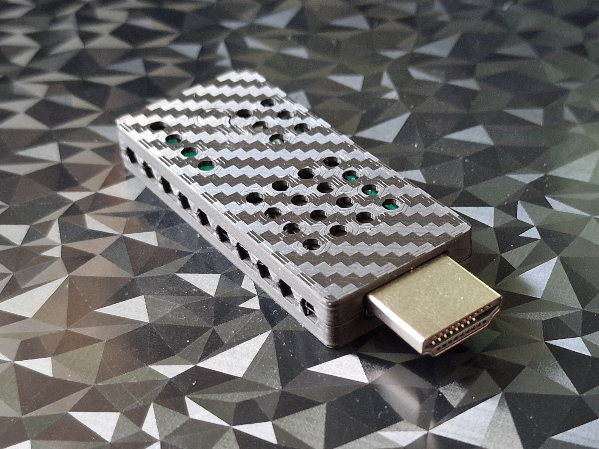 Szary adapter HDMI z wzorem w jodełkę i otworami wentylacyjnymi, leżący na błyszczącym, geometrycznym tle. Makro, ostre detale.