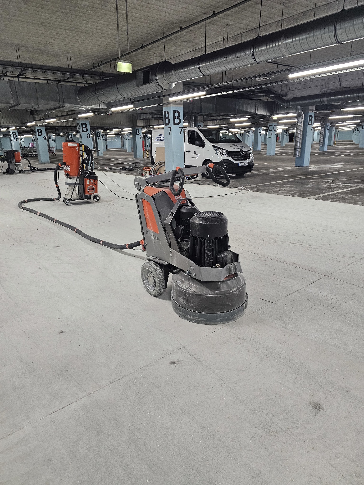 Szlifowanie betonowej posadzki w garażu podziemnym. Szlifierka Husqvarna PG 680 RC z odkurzaczem przemysłowym. W tle biały bus i słupy z oznaczeniami.