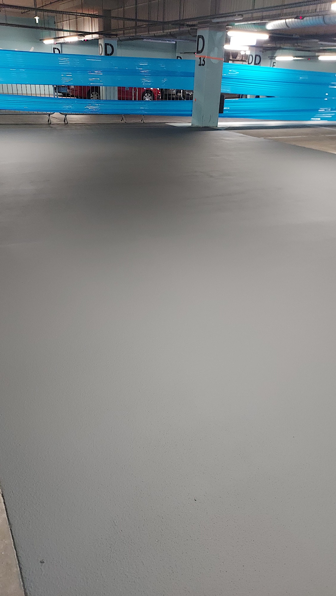 Szara, gładka posadzka w garażu podziemnym, zabezpieczona niebieską taśmą. Widoczne słupy z oznaczeniami stref parkowania. Minimalistyczny design, świeżo po wylaniu.