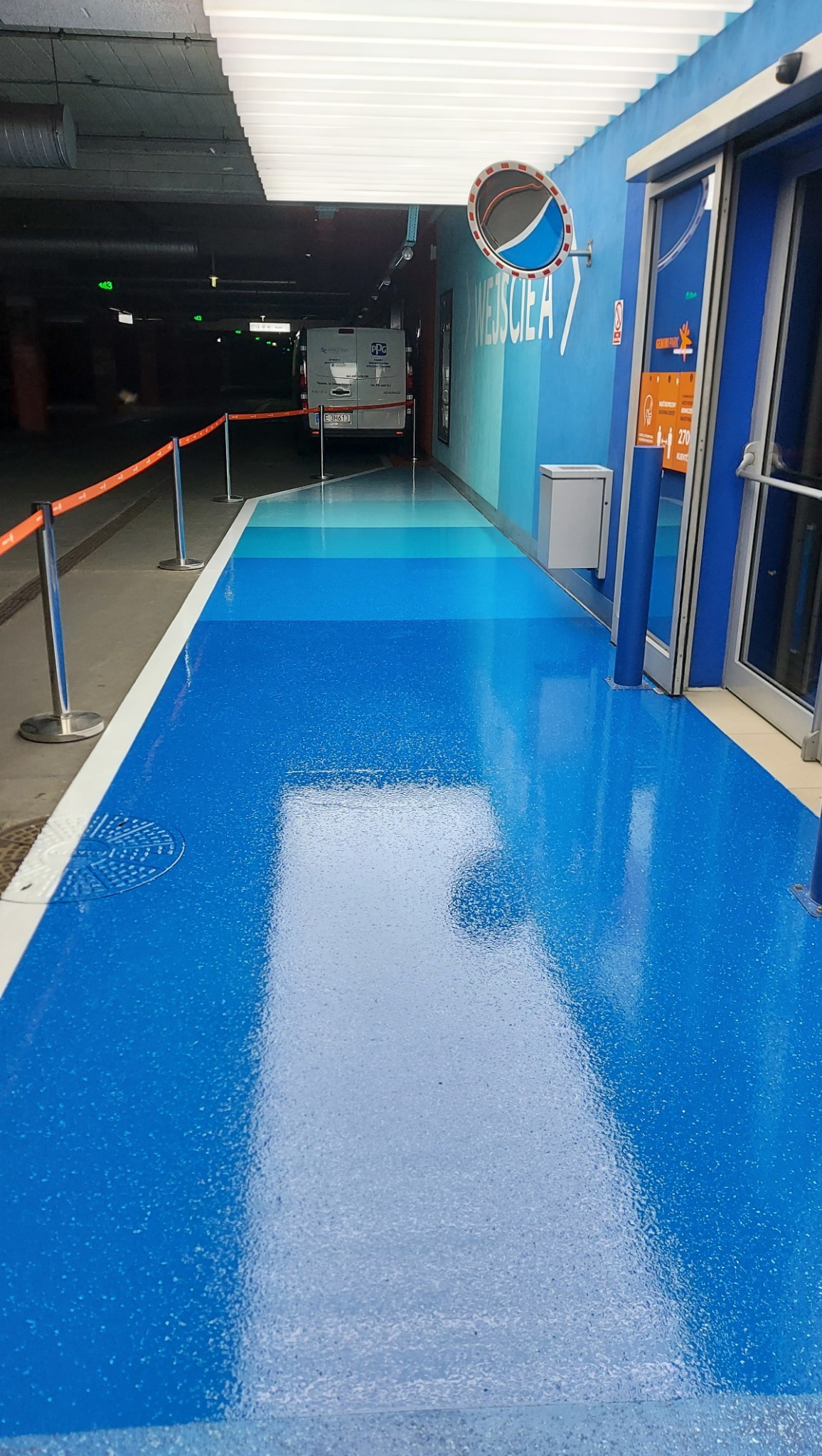 Błękitna posadzka żywiczna w garażu podziemnym, z widocznym gradientem kolorów i oznaczeniami kierunkowymi. W tle zaparkowany bus.