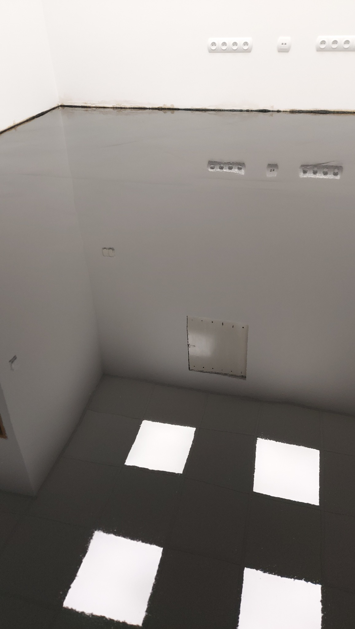 Błyszcząca, szara posadzka w nowym pomieszczeniu z białymi ścianami i widocznymi gniazdkami elektrycznymi. Na podłodze widoczne odbicia światła.
