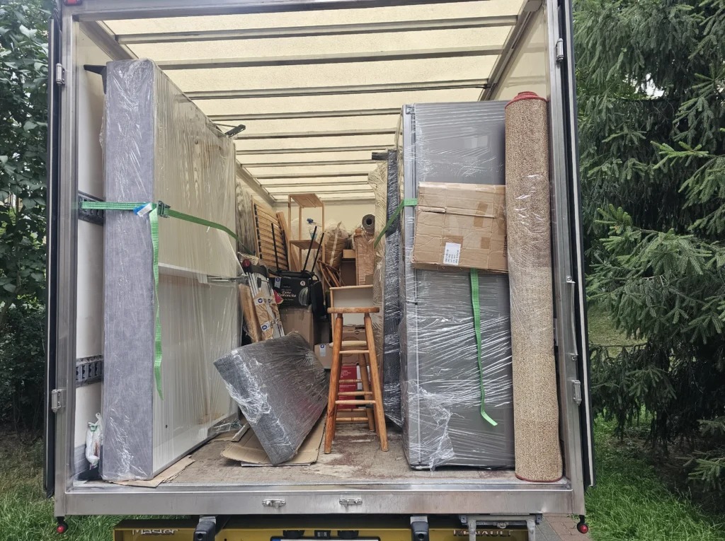 Wnętrze furgonetki wypełnionej po brzegi meblami i sprzętami, zabezpieczonymi folią i pasami transportowymi, gotowymi do przewozu. Widok z tyłu pojazdu.