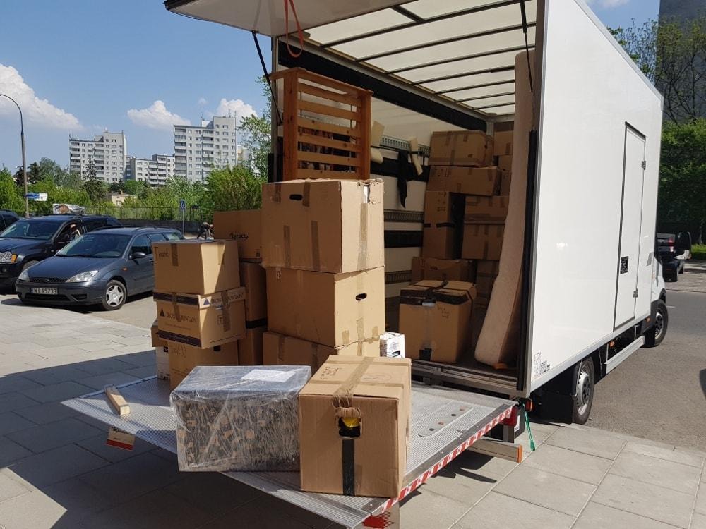 Otwarty samochód dostawczy wypełniony kartonami i meblami podczas przeprowadzki na tle miejskiego krajobrazu z budynkami mieszkalnymi i zaparkowanymi autami.