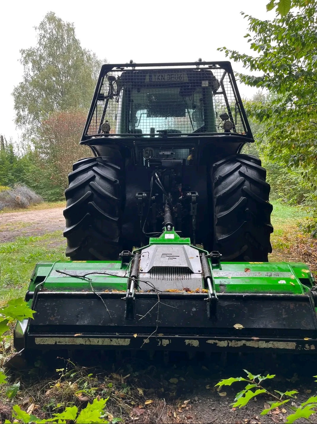 Ciężki traktor z mulczerem leśnym w lesie. Widok z tyłu na maszynę gotową do pracy. Zielony osprzęt, duże opony. TKN 3EU61 na tablicy rejestracyjnej.