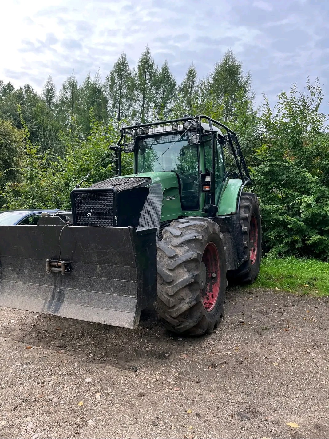Zielony traktor z czarnym pługiem na tle drzew. Sprzęt ciężki do prac leśnych lub ziemnych. Czerwone felgi. Pochmurne niebo.