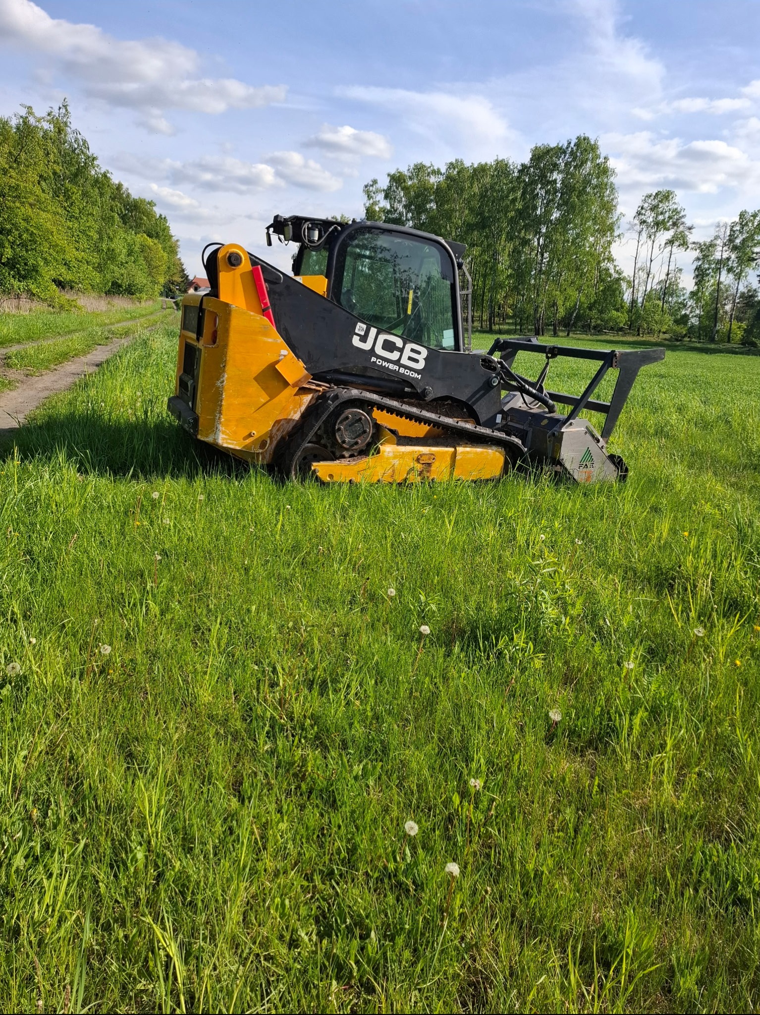 Żółto-czarna kosiarka trawnikowa JCB na gąsienicach, z widoczną głowicą tnącą, na tle zielonej trawy i drzew pod błękitnym niebem z chmurami.