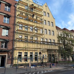 Żółta kamienica w trakcie renowacji, otoczona rusztowaniami. Pracownicy na różnych poziomach rusztowań. Widoczne detale architektoniczne i sąsiednie budynki.
