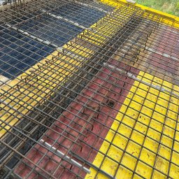 Zbrojenie stropu budynku w trakcie budowy, widok z góry. Żebrowana siatka stalowa na deskowaniu z żółtymi i czerwonymi elementami. Widoczne pręty pionowe.